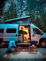 Peaceful RV rental scene: illuminated van, cozy chairs, and warm lights under towering trees at dusk.