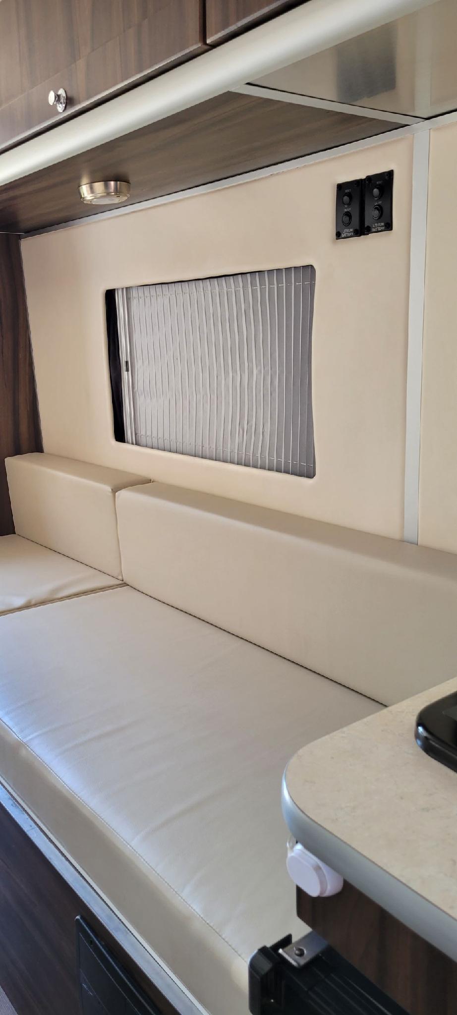 Light-colored upholstered bench seating and window with pleated blind inside an RV rental. Overhead cabinets and battery controls visible. — photo 7