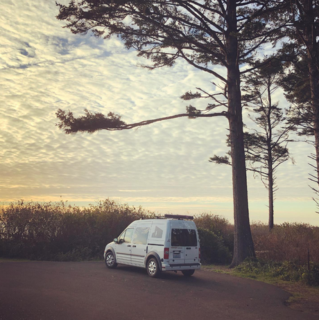 White rv rental van parked peacefully by tall trees on a dirt road, offering coastal views under a beautiful cloudy sky.