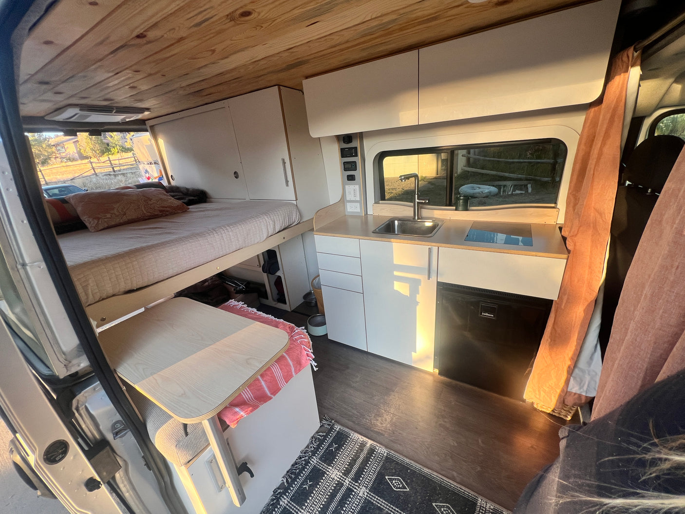 This inviting RV rental offers a raised bed, modern kitchenette with sink & fridge, and controls for an amazing van life.