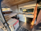 This inviting RV rental offers a raised bed, modern kitchenette with sink & fridge, and controls for an amazing van life.