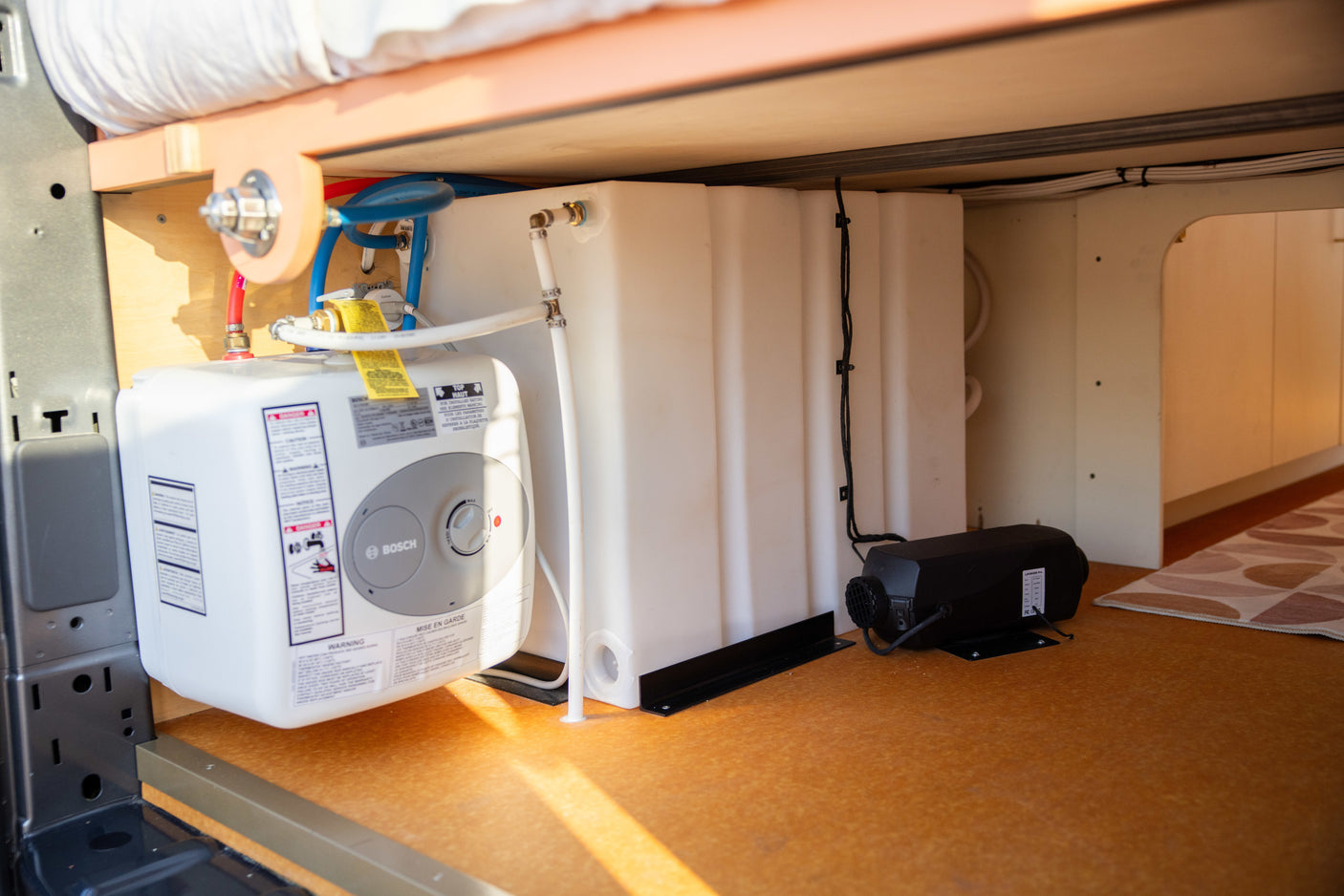 RV rental: Under-bed view of a Bosch water heater, fresh water tank, and diesel heater with plumbing.