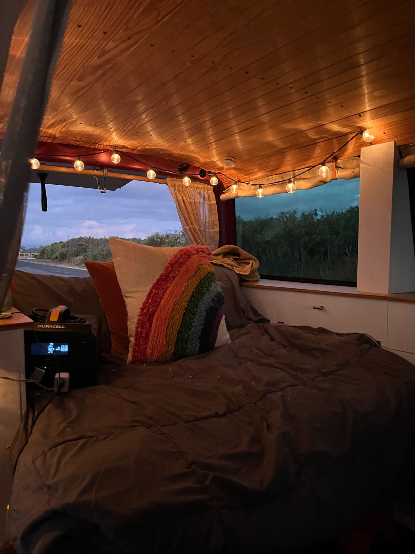 Inside a cozy RV rental: warm string lights, a comfy bed, and a power station with a tranquil evening view.