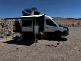 RV rental van life setup: Sprinter van with awning, roof tent, people & dog relaxing in a sunny, arid landscape.