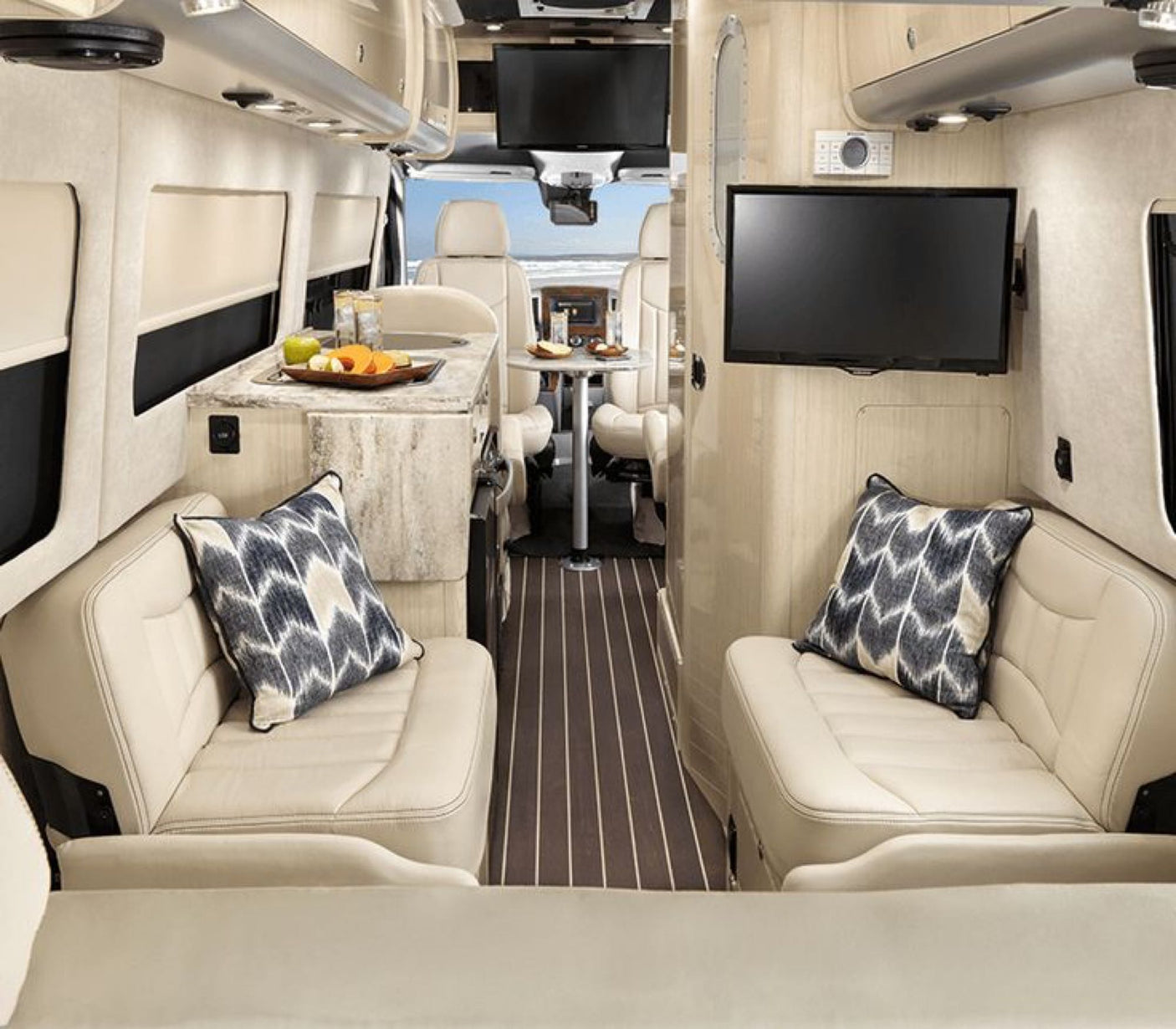 This luxury RV rental interior features comfy seating, a kitchen, TV, and a dining area, with a beach view through the front.