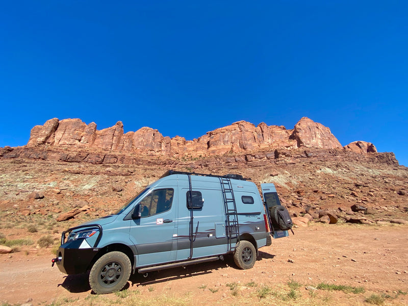 This rugged RV rental camper van is parked on a desert road, with towering red rock formations under a clear blue sky. — photo 9