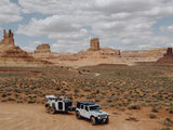 White Jeep and overland RV rental trailer on a dirt road in a vast desert landscape with imposing rock formations under a cloudy sky.