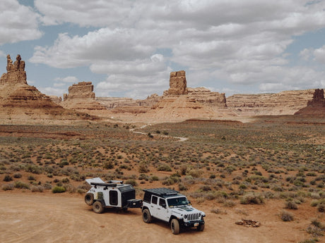 White Jeep and overland RV rental trailer on a dirt road in a vast desert landscape with imposing rock formations under a cloudy sky.