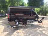 RV rental camper van at a sunny campsite. Awning extended over outdoor chairs and table on a mat for relaxing.