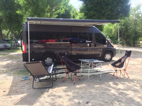 RV rental camper van at a sunny campsite. Awning extended over outdoor chairs and table on a mat for relaxing.