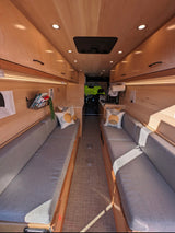 Modern RV rental interior: Light wood walls, overhead cabinets, and two long grey cushioned benches create a cozy living space.