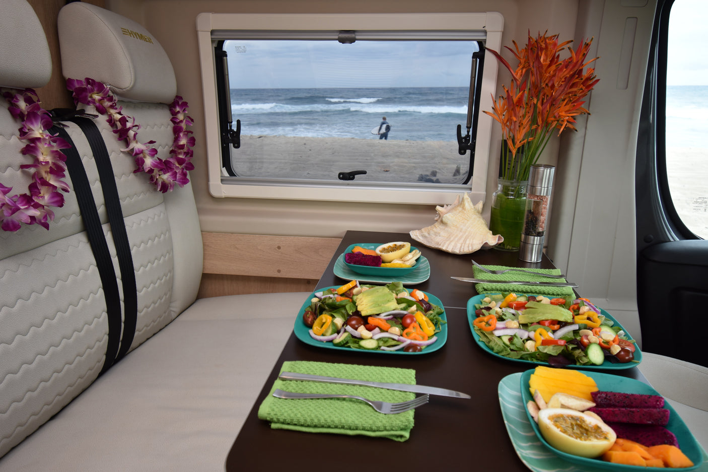 RV rental dining: Fresh salads, fruit, and leis. Enjoy the beach view with a surfer and waves from inside the camper.