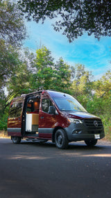 Maroon Mercedes Sprinter RV rental parked on a road, side door open to reveal a custom interior, surrounded by green trees.