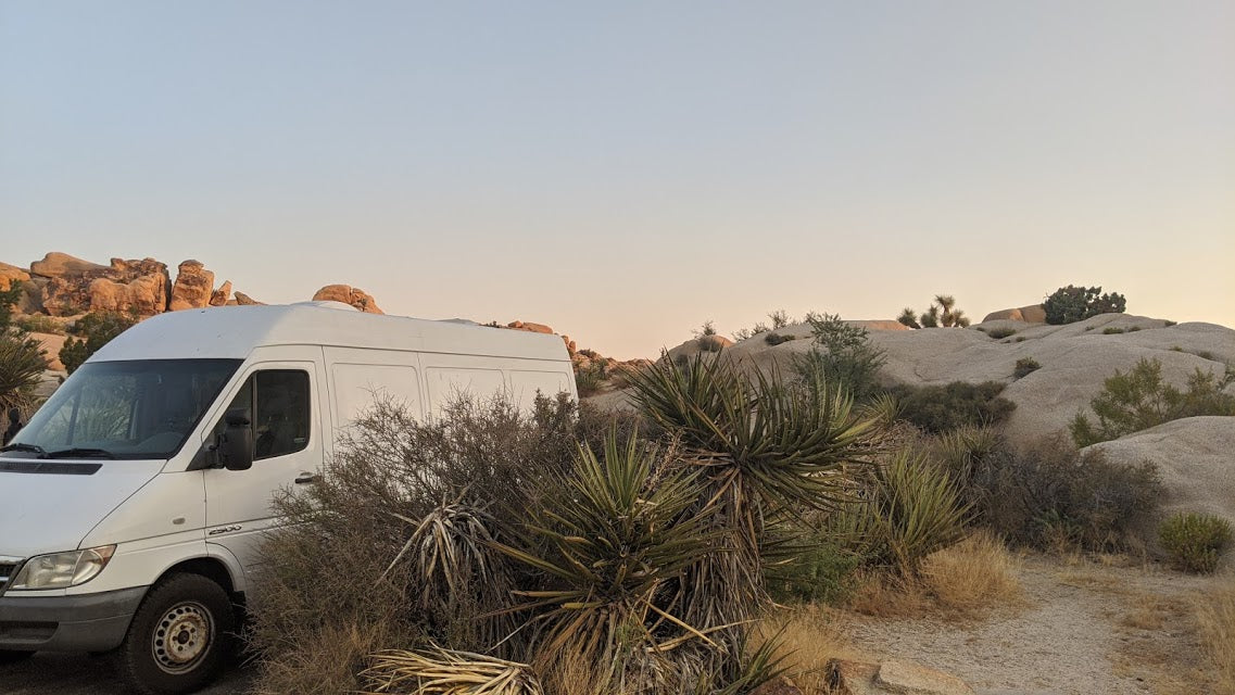 White RV rental van nestled in a desert landscape with large boulders and yuccas at dusk.
