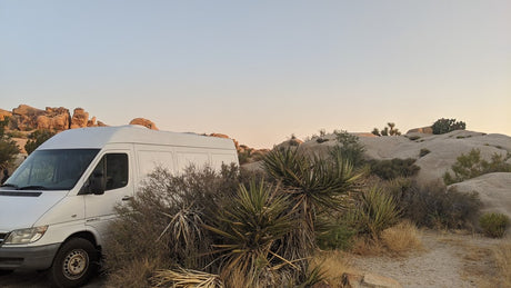 White RV rental van nestled in a desert landscape with large boulders and yuccas at dusk.