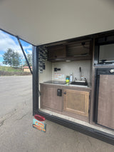 This RV rental's outdoor kitchen features a sink, cooktop, microwave, and fridge with wood-grain cabinets and a tiled backsplash.