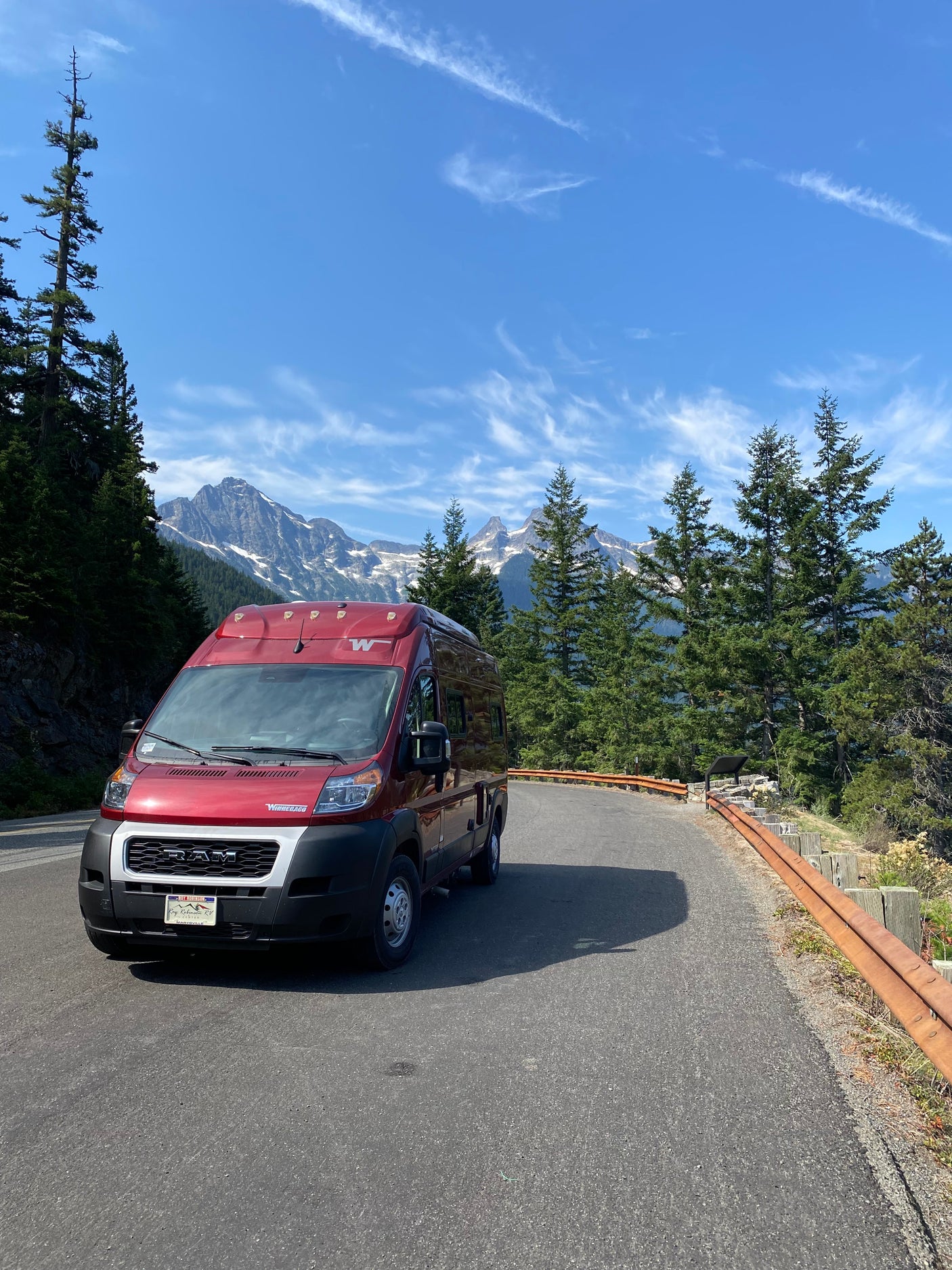 Red rv rental on a scenic mountain road, surrounded by tall trees and snow-capped peaks under a blue sky.