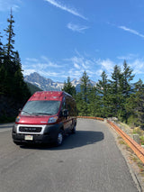 Red rv rental on a scenic mountain road, surrounded by tall trees and snow-capped peaks under a blue sky.