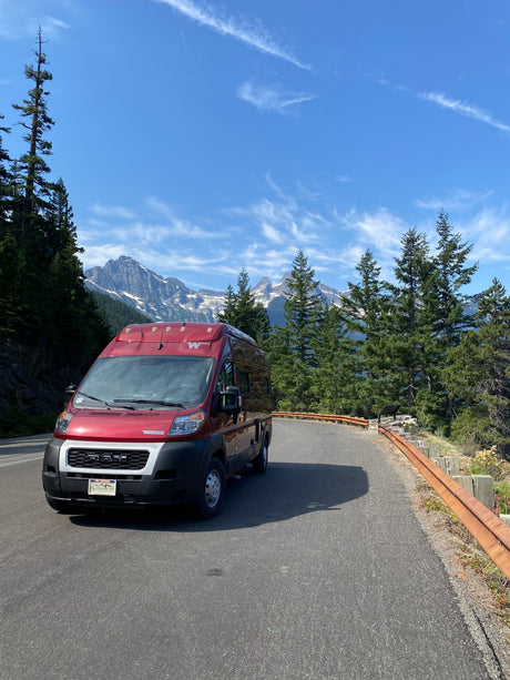 Red rv rental on a scenic mountain road, surrounded by tall trees and snow-capped peaks under a blue sky.