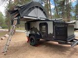 Off-road camper trailer RV rental with rooftop tent, set up for camping among pine trees. Ideal for outdoor adventures.