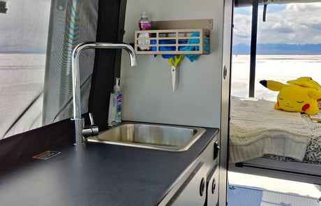 RV rental interior featuring a practical kitchenette with a sink & storage, plus a comfy bed with Pikachu overlooking a vast, scenic landscape.