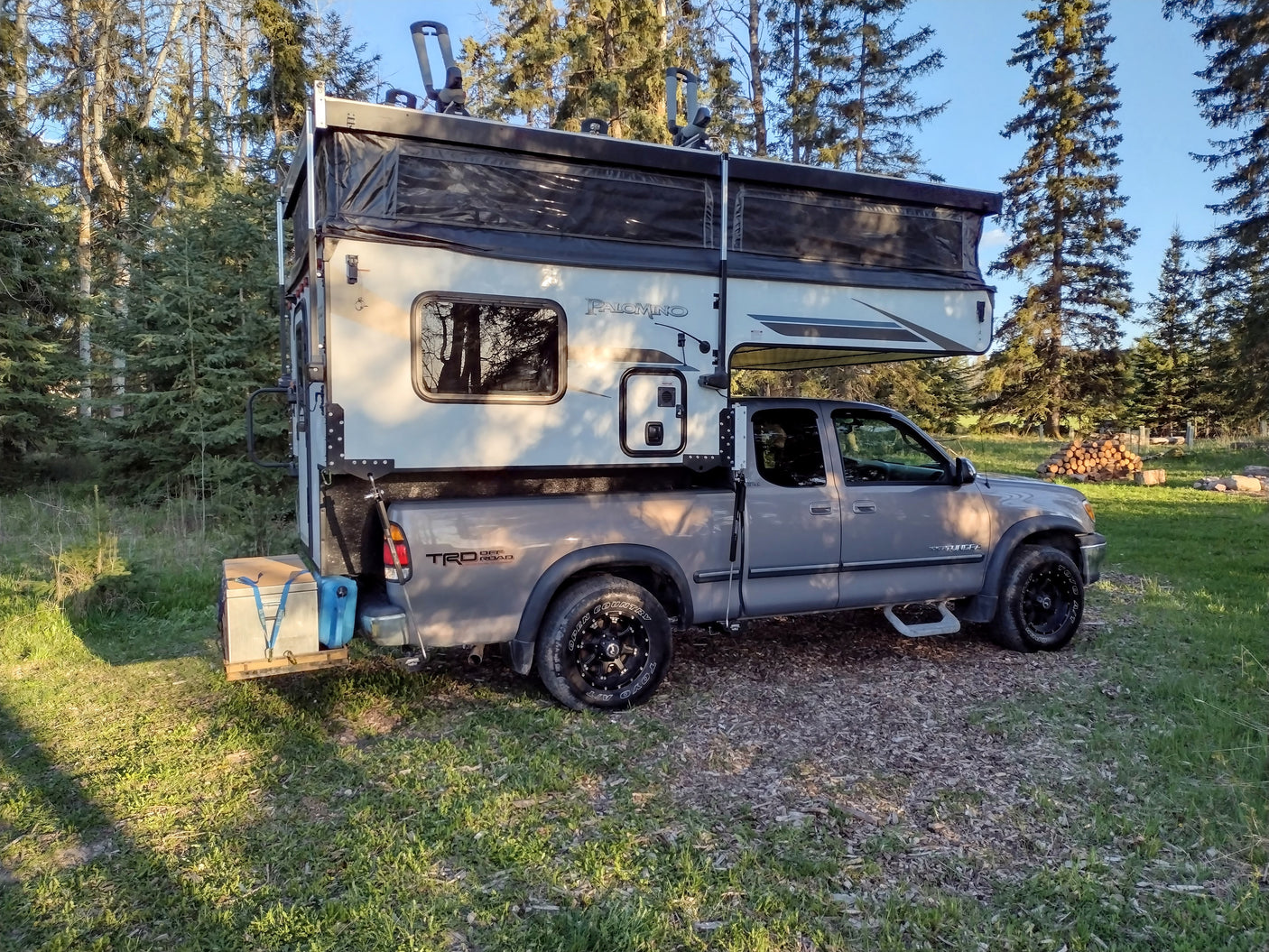 Rugged grey pickup with a pop-up truck camper parked in a wooded area. An excellent RV rental for off-road camping.