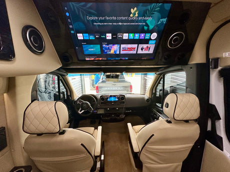 Luxurious RV rental interior, featuring a large overhead Smart TV for entertainment, quilted cream leather seats, and a Mercedes-Benz cockpit.