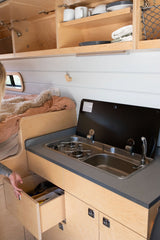 Inside a compact RV rental, showcasing a kitchen with a stove, sink, open utensil drawer, and overhead cabinets next to a bed.