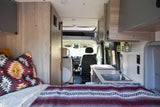 Inside a modern RV rental: a cozy bed with patterned blanket, compact kitchen sink, and view to the driver's cabin.