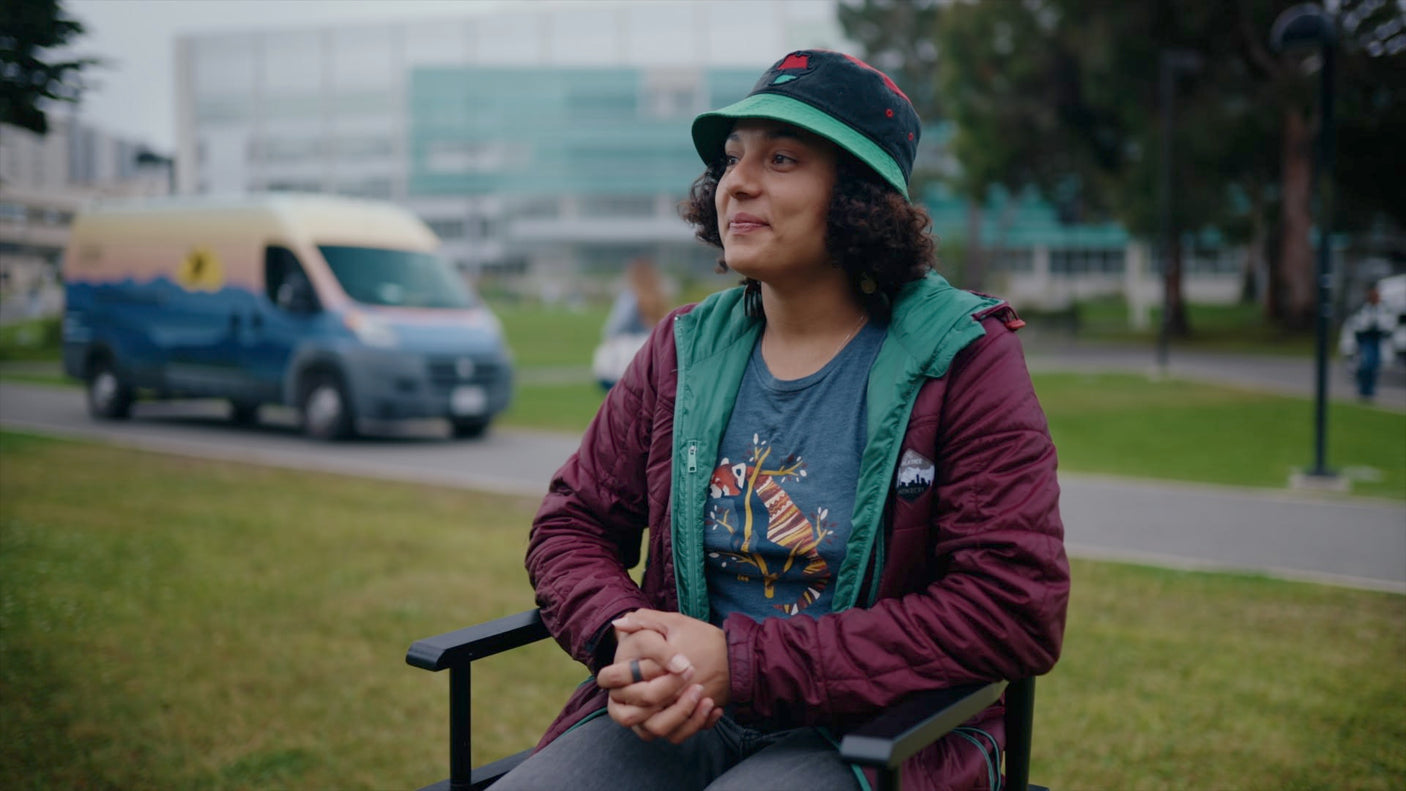A smiling person in a hat and jacket sits outdoors, hands clasped. A colorful rv rental van is blurred behind them.