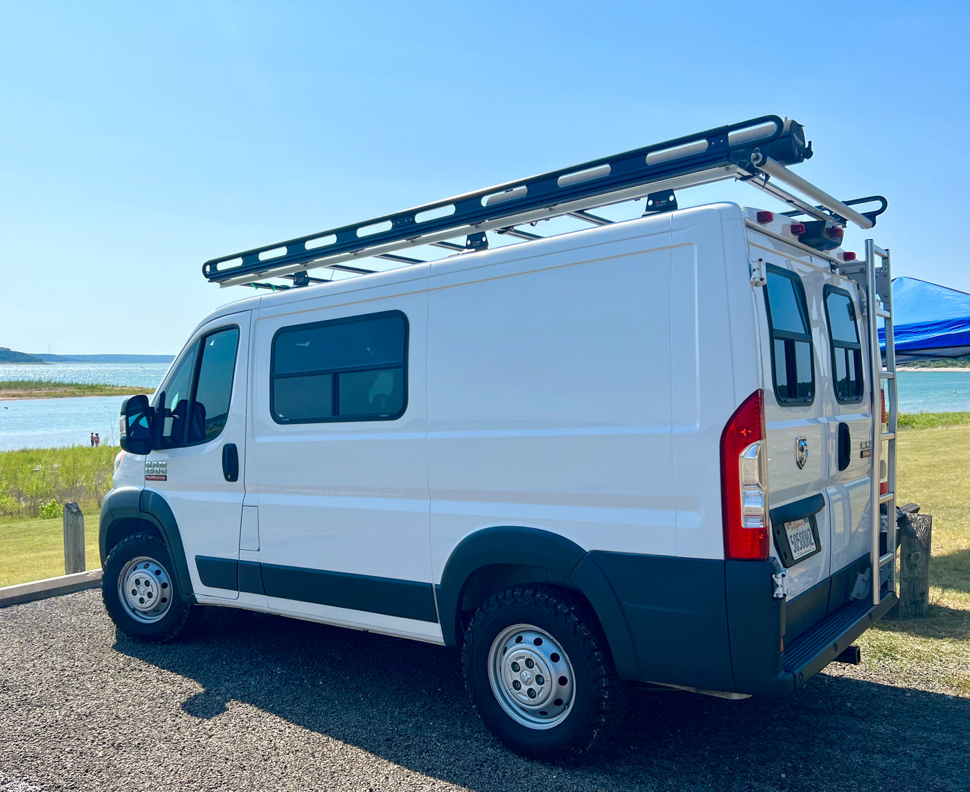 A white Ram ProMaster camper van with a roof rack and ladder, parked by a sunny lake. Ideal for your next rv rental adventure. — photo 14