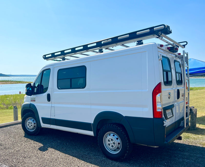 A white Ram ProMaster camper van with a roof rack and ladder, parked by a sunny lake. Ideal for your next rv rental adventure. — photo 14