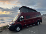 Maroon pop-top camper van, an ideal rv rental, parked by the ocean at dusk. Clouds glow as waves meet the shore.