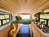 Cozy RV rental interior: light wood ceiling, elevated bed with green comforter, compact kitchen with sink & stove.