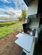 Scenic RV rental view: a camper van kitchen with stove/sink opens to lush green land and a beautiful ocean vista.