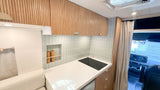 Bright RV rental galley: light wood slatted cabinets, pale green vertical subway tile backsplash, white counter with cooktop.