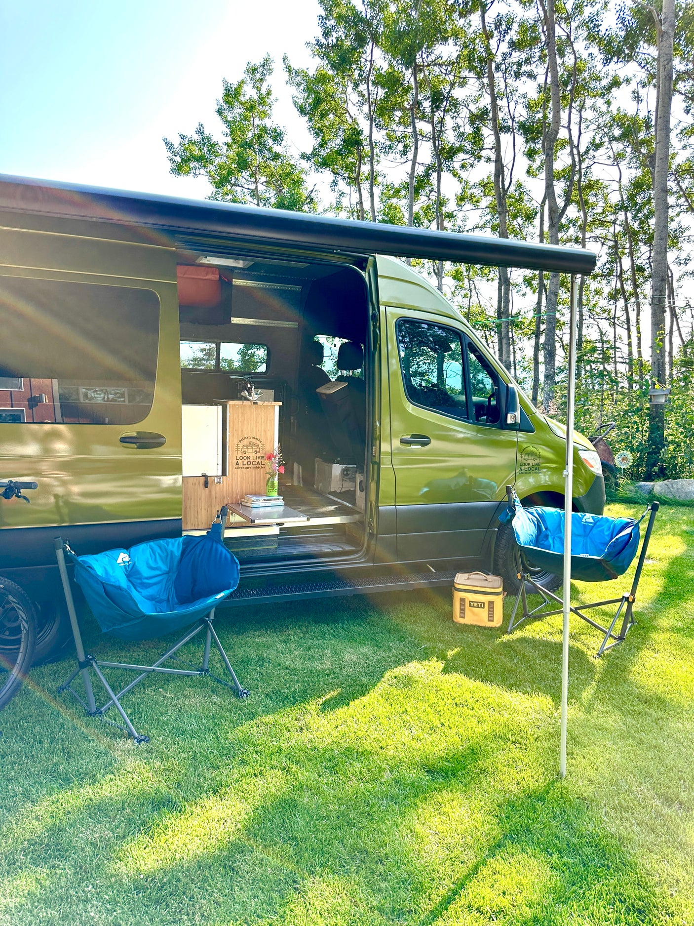 Green adventure RV rental parked on grass with awning extended, blue chairs, and a Yeti cooler. Perfect for outdoor relaxation.