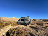 This grey Storyteller Overland 4x4 adventure van, ideal for an RV rental, is parked on a desert hill with arid mountains.