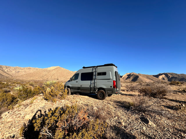 This grey Storyteller Overland 4x4 adventure van, ideal for an RV rental, is parked on a desert hill with arid mountains.