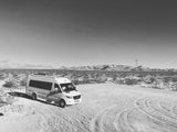 B&W photo of a white Mercedes Sprinter RV rental parked in a vast desert with distant mountains & a power tower.