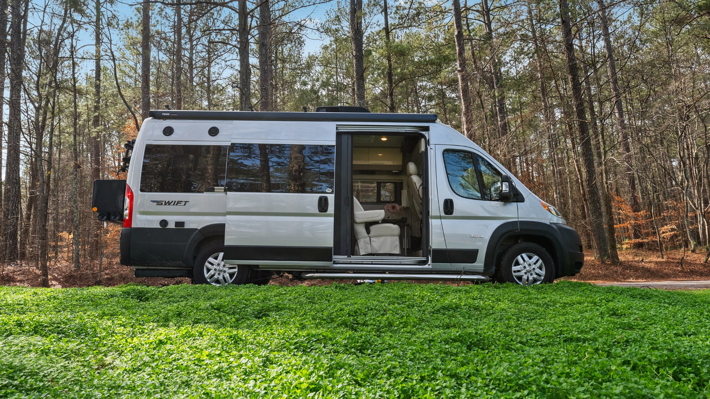 This Class B RV rental, a Swift camper van, is parked amidst lush green ground with a forest backdrop. The side door is open, revealing its cozy interior.
