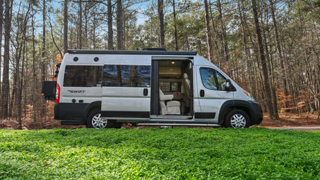 This Class B RV rental, a Swift camper van, is parked amidst lush green ground with a forest backdrop. The side door is open, revealing its cozy interior.