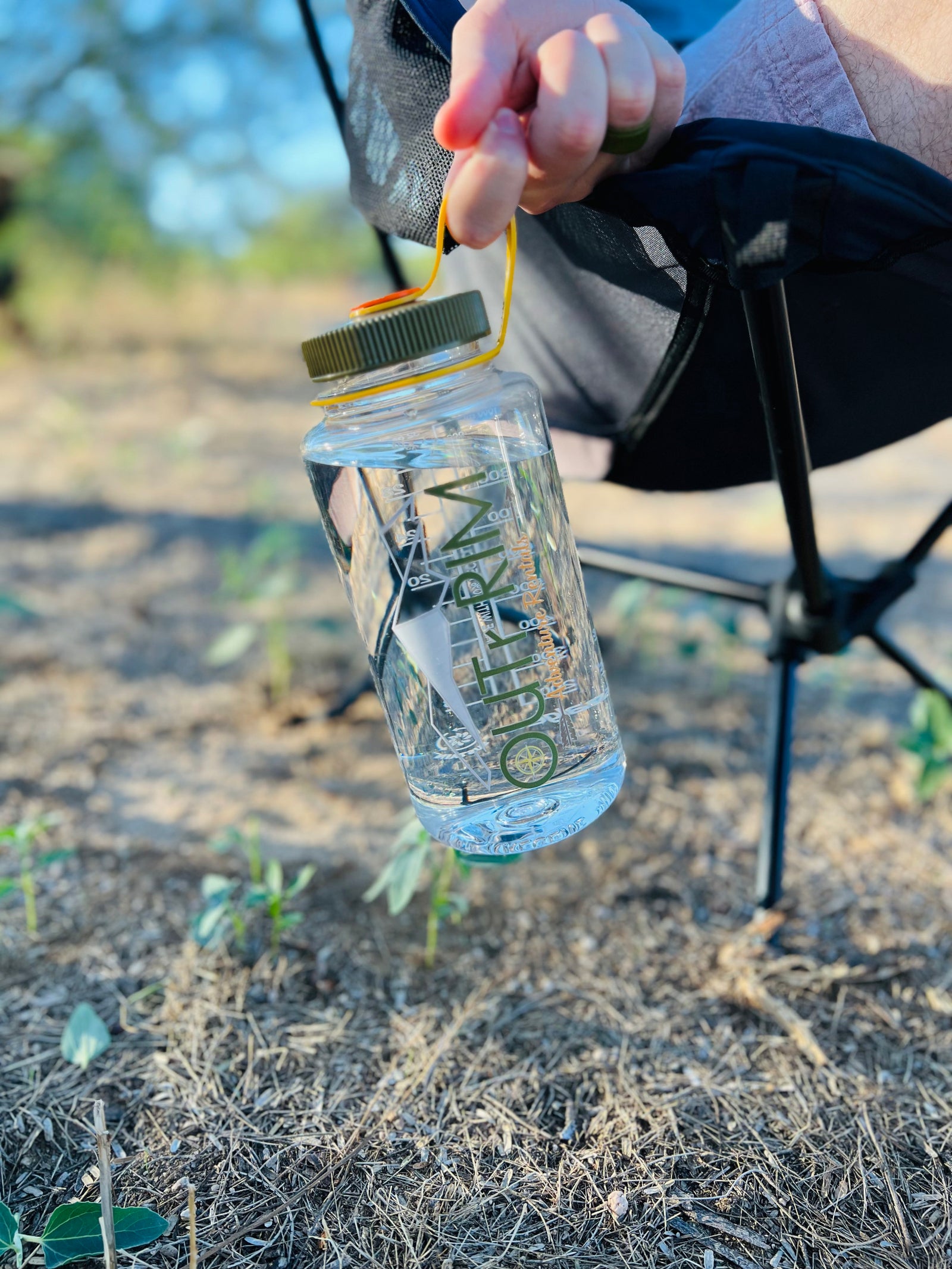 Enjoying an RV rental trip! A person holds a clear water bottle with "OUTRKM Adventure Rentals" branding, seated outdoors.