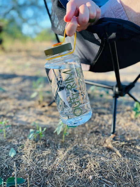 Enjoying an RV rental trip! A person holds a clear water bottle with "OUTRKM Adventure Rentals" branding, seated outdoors.