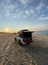 White truck camper on sandy beach at sunset, surfboards on top. Your next RV rental adventure awaits!