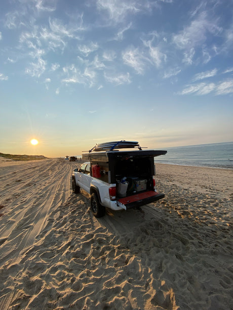 White truck camper on sandy beach at sunset, surfboards on top. Your next RV rental adventure awaits!