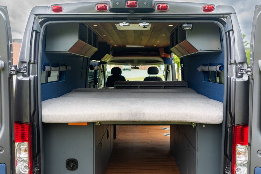 This RV rental showcases a camper van's interior with a comfy bed, wood floor storage, and overhead cabinets for adventure.