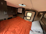 This rv rental shows a cozy bedroom with an orange bed, AC unit, thermostat, and storage, perfect for your next trip.