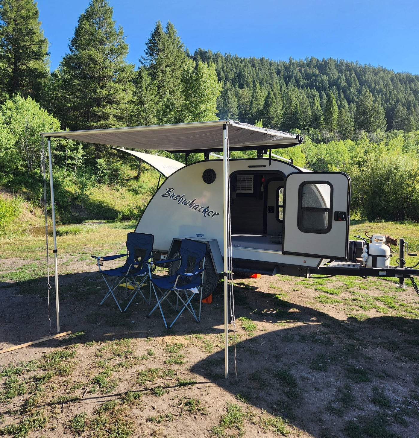 Bushwhacker RV rental set up in a scenic forest clearing with awning, chairs, and a dog, ready for outdoor adventure. — photo 9