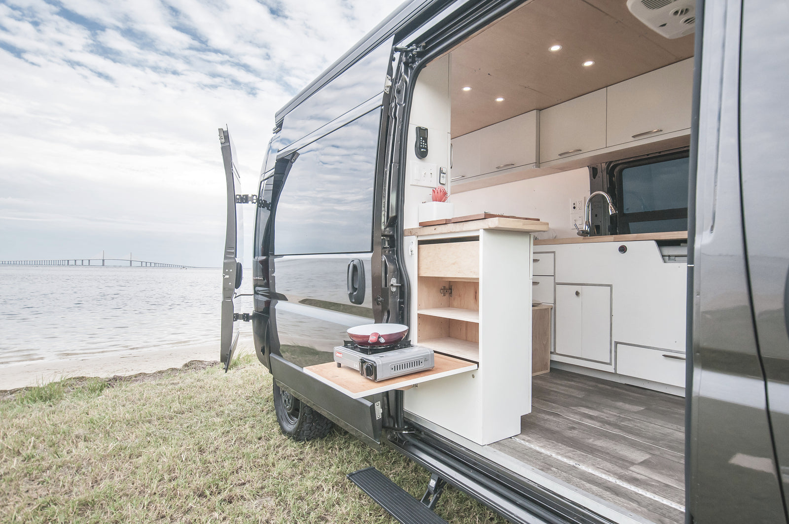 Experience outdoor cooking with this RV rental camper van's pull-out kitchen, parked by a bay with a distant bridge.